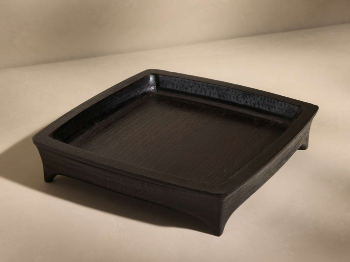 Square black tray with a slightly raised edge, made of textured wood, resting on a light beige surface, viewed at an angle.