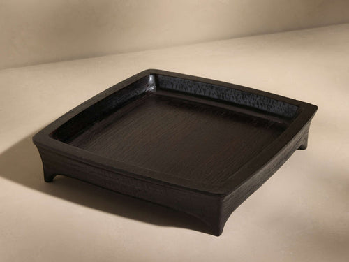 Square black tray with a slightly raised edge, made of textured wood, resting on a light beige surface, viewed at an angle.