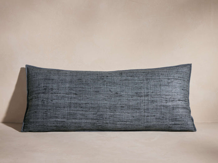 Horizontal dark blue-grey Raw Silk Pillow with visible textured weave and merrow stitch detail, casting shadow on a neutral background.