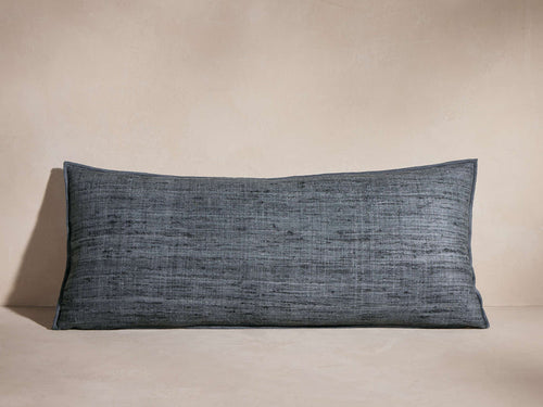 Horizontal dark blue-grey Raw Silk Pillow with visible textured weave and merrow stitch detail, casting shadow on a neutral background.