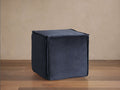 Dark blue washed velvet cubic pouf with visible flanged seams sits on a wooden floor against a beige wall.