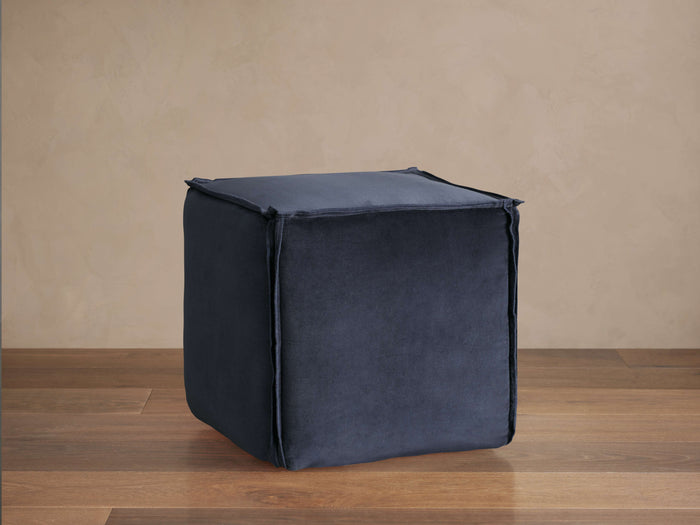 Dark blue washed velvet cubic pouf with visible flanged seams sits on a wooden floor against a beige wall.