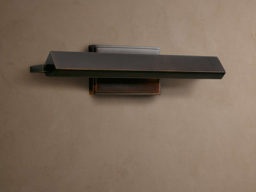 Zaeden Picture Light, dark bronze-plated with a horizontal angled bar and rectangular backplate, mounted on a textured brown wall.
