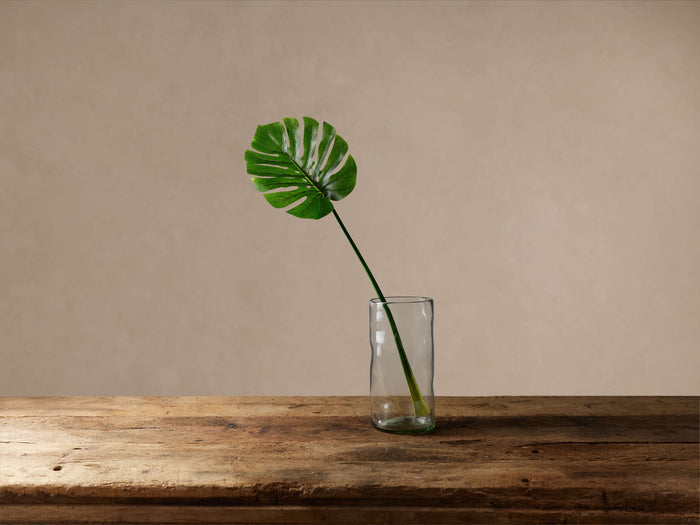 A vibrant green faux split leaf philodendron stem in a clear glass vase on a rustic wooden table, beige wall background.