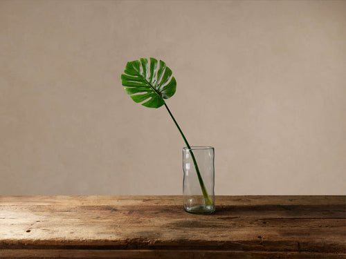 A vibrant green faux split leaf philodendron stem in a clear glass vase on a rustic wooden table, beige wall background.