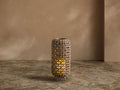 Noale Outdoor Lantern, cylindrical light brown handwoven wicker with a warm glowing LED candle, on stone floor.
