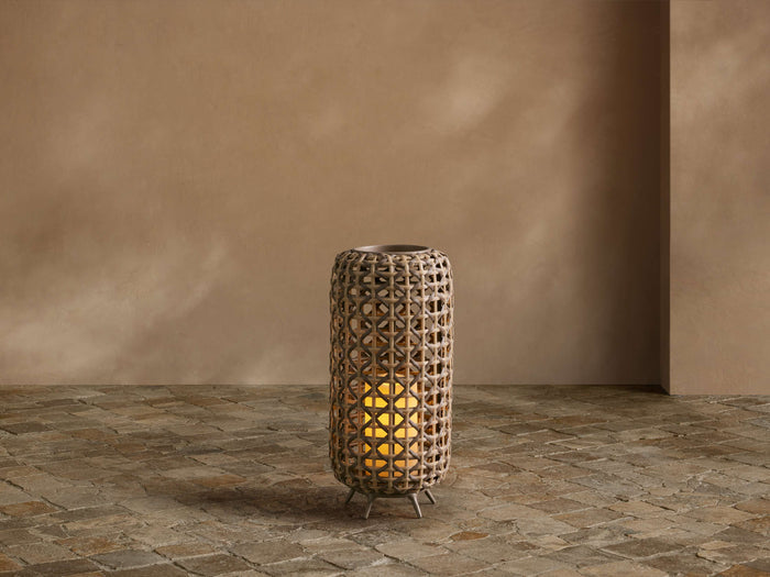 Noale Outdoor Lantern, cylindrical light brown handwoven wicker with a warm glowing LED candle, on stone floor.