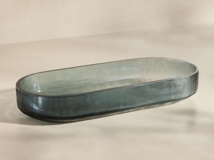 Front view of a smoky gray translucent oval glass tray with fine linear texture, shown from an elevated angle.
