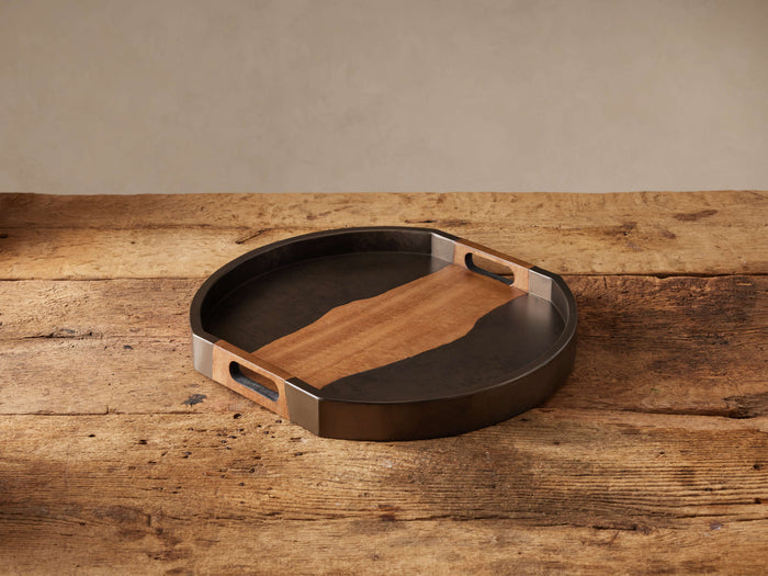 Acacius Round Tray: dark oxidized steel frame, warm brown yukas wood inlay with organic edge and handles, on rustic wood.