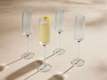 Four Ada reeded champagne flutes on a light surface. One glass is filled with sparkling yellow liquid & lemon peel.