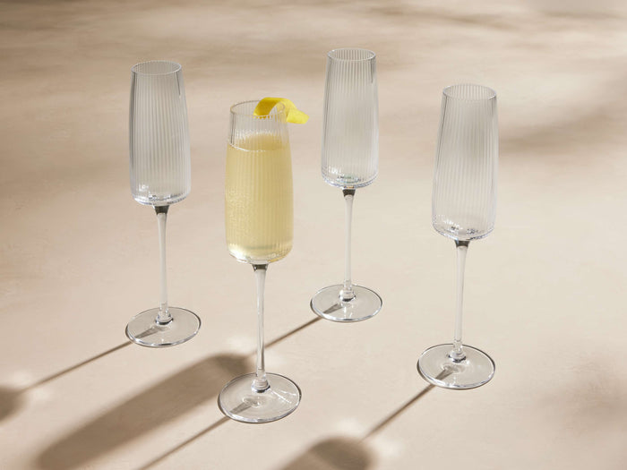 Four Ada reeded champagne flutes on a light surface. One glass is filled with sparkling yellow liquid & lemon peel.