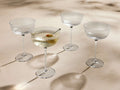 Four clear reeded Ada Coupe Glasses, one holding a martini with an olive, arranged on a light surface with shadows.
