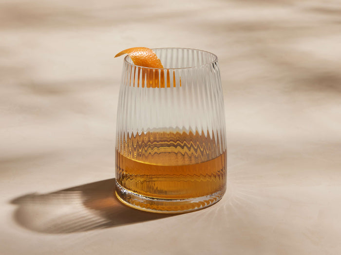 Ada Double Old Fashioned Glass, clear, reeded texture, filled with amber liquid, garnished with orange peel, on a light surface.