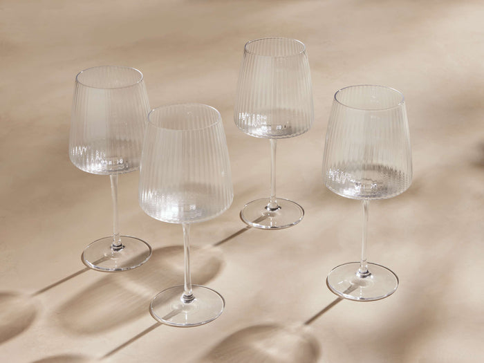 Set of four Ada Wine Glasses with reeded design, clear glass, wide bowls on elegant stems, arranged on a soft beige surface.