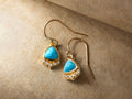Adira Turquoise Earrings with 8 mm x 10 mm blue turquoise drops set in gold vermeil, accented with small round crystals, on a tan surface.