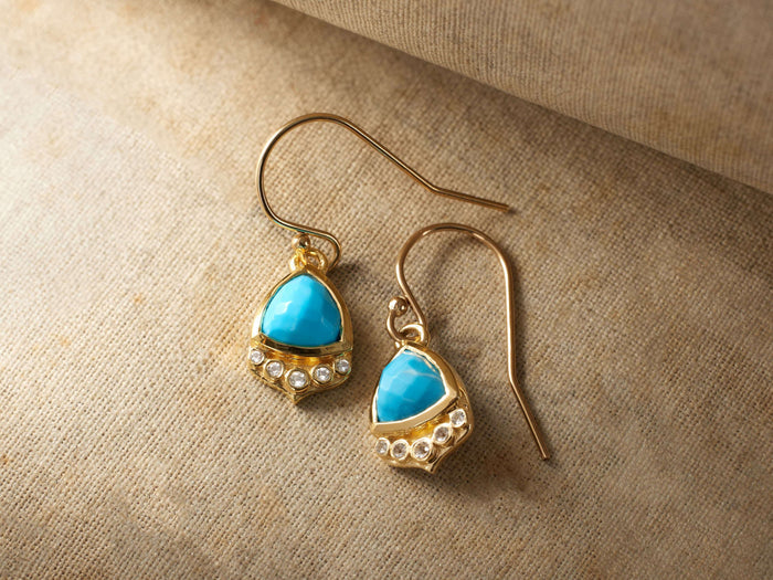 Adira Turquoise Earrings with 8 mm x 10 mm blue turquoise drops set in gold vermeil, accented with small round crystals, on a tan surface.