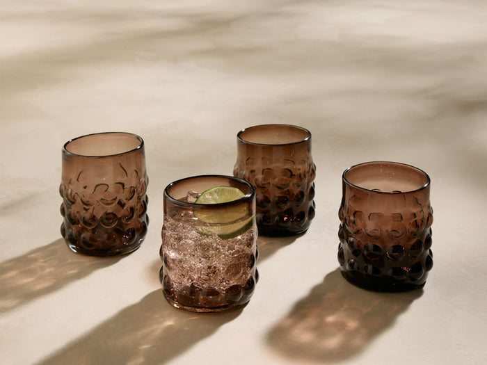 Four amber Aera Double Old Fashioned Glasses with a bubbly texture. One glass contains ice and a lime slice.