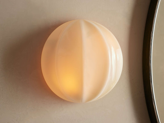 Agnes Clamshell Sconce: Spherical cream paneled glass sconce, glowing warmly, mounted on a light wall next to a dark mirror.