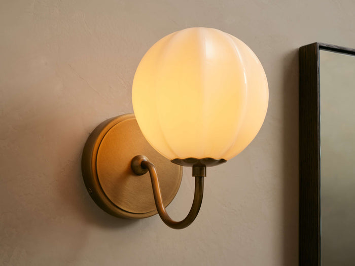 Agnes Gooseneck Sconce with antique brass base, curved arm, and glowing paneled cream glass globe shade mounted on a wall.