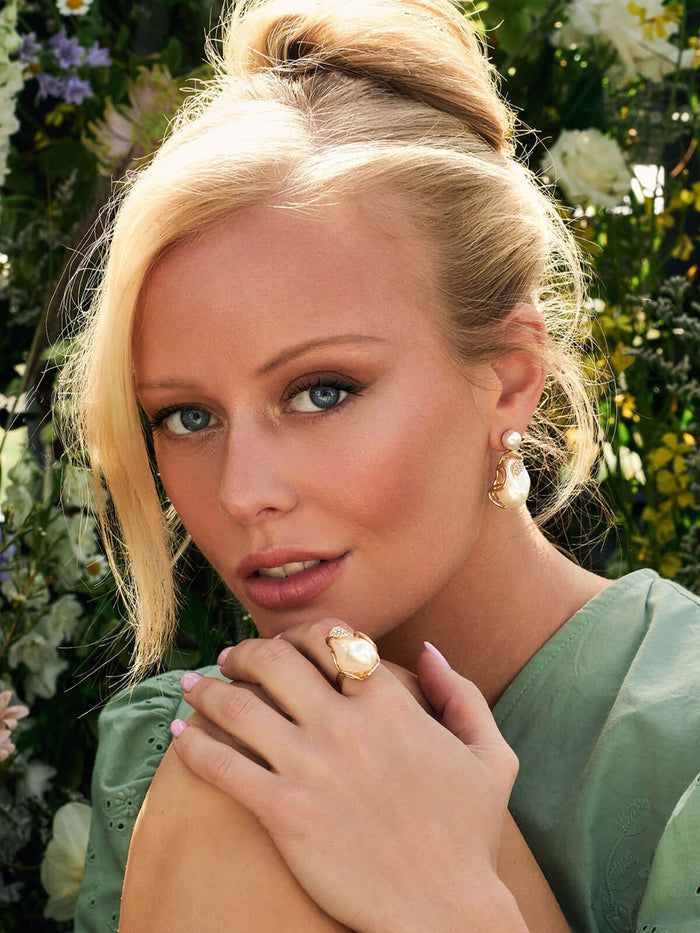 Woman with blonde hair and green eyes wearing a gold ring, surrounded by flowers.
