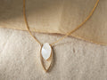 Allaire Necklace on a beige fabric, featuring a marquise-cut mother of pearl gemstone within a gold fill lotus petal pendant on a gold chain.