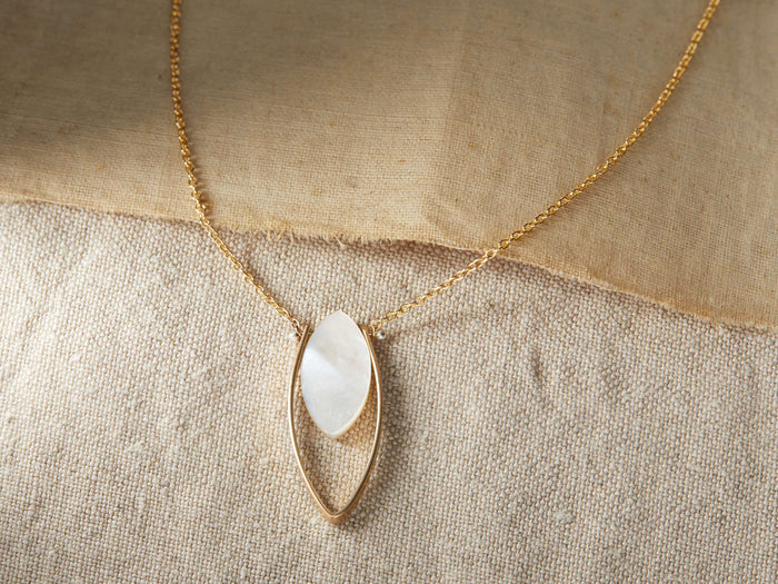 Allaire Necklace on a beige fabric, featuring a marquise-cut mother of pearl gemstone within a gold fill lotus petal pendant on a gold chain.
