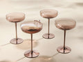 Front view of four Ambretta Coupe Glasses with mocha-smoke glass bowls and metallic bronze stems and bases. One holds a drink with lavender.