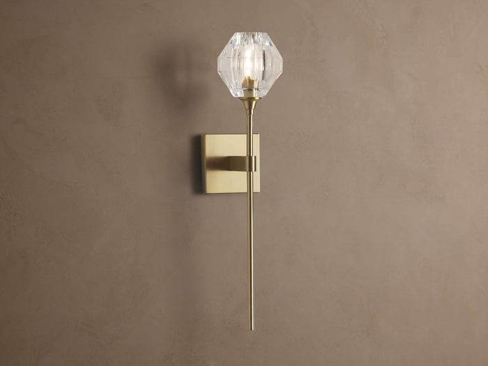 Amoroso Sconce: brass-plated wall light with long rod and illuminated faceted clear crystal shade on a brown wall.