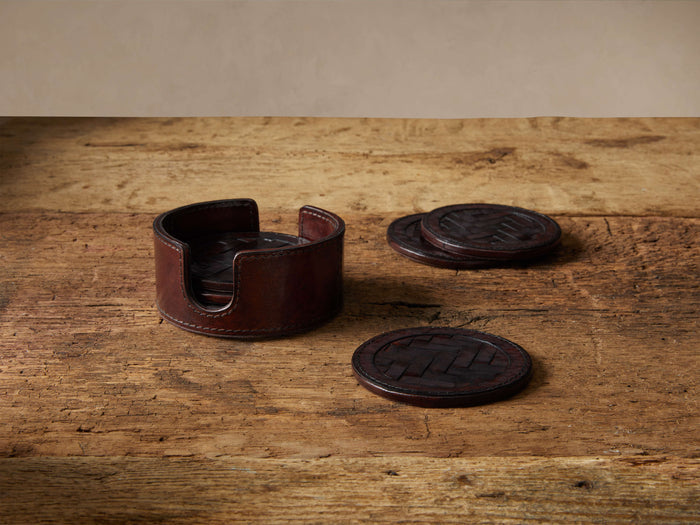 Dark brown Andres Woven Coasters set on a rustic wooden table. Leather holder, stacked & single woven coasters visible.