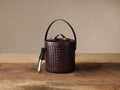 Andres Woven Ice Bucket: Dark brown woven leather bucket with matching lid, arched handle, and brass accents, on wood. Tongs hang from side.