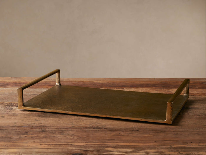 Angelo Rectangular Tray in antique brass aluminum, shown at an angle on a wooden surface, with raised, rectangular handles.