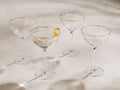 Four Anisa ribbed cocktail glasses with gold rims, one filled with liquid & lemon peel, on a light surface.