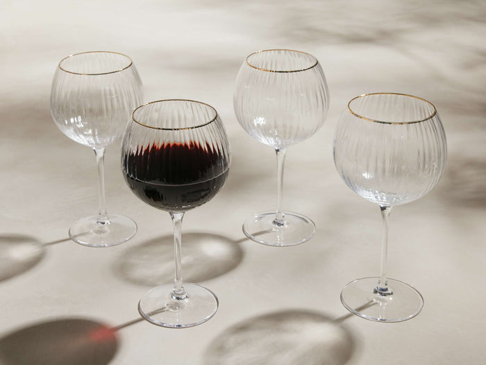 Front view of four clear Anisa Red Wine Glasses with ribbed bowls and gold rims, one filled with red wine.