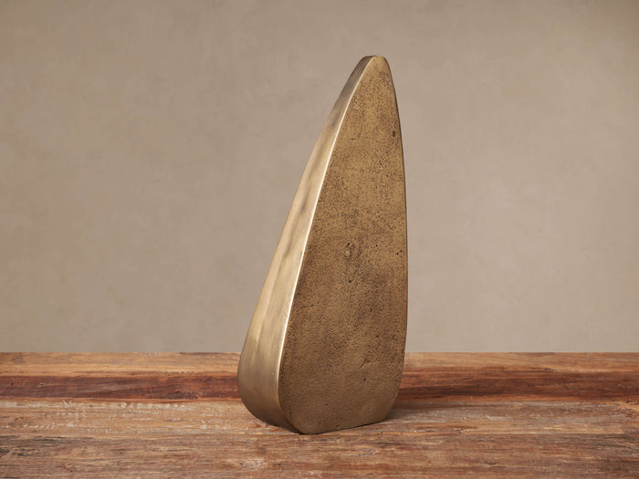 Abstract, curved antique brass sculpture with textured finish stands on rustic wood surface.