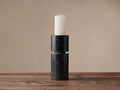 Black marble Aphra Pillar candle holder, two stacked sections with white veins, holding an ivory pillar candle with a lit flame on a wooden table.
