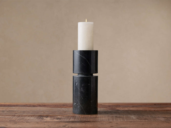 Black marble Aphra Pillar candle holder, two stacked sections with white veins, holding an ivory pillar candle with a lit flame on a wooden table.