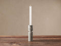 Aphra Tall Taper Holder, a two-piece light gray marble holder with dark veins, holds a lit white taper candle on a wood surface.