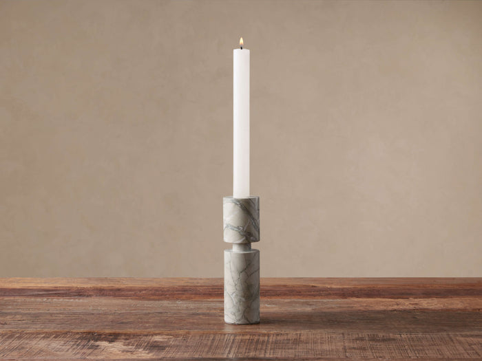Aphra Tall Taper Holder, a two-piece light gray marble holder with dark veins, holds a lit white taper candle on a wood surface.