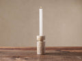 Aphra Taper Holder, a cylindrical natural beige marble candle holder with dark veins, holding a tall, lit white taper candle.