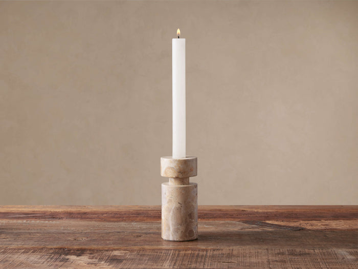 Aphra Taper Holder, a cylindrical natural beige marble candle holder with dark veins, holding a tall, lit white taper candle.