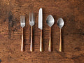 Arber Five Piece Place Setting in Copper on a textured wooden surface. Includes two forks, a knife, and two spoons with distressed handles and polished tops.