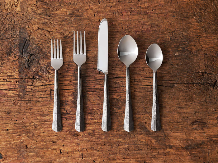Five-piece Arber place setting on wood surface, featuring two forks, a knife, and two spoons with blackened handles and polished tops.
