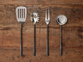 Set of 4 Arber Kitchen Tools: silver polished slotted spatula, pasta server, carving fork, and ladle with dark handles on wood.