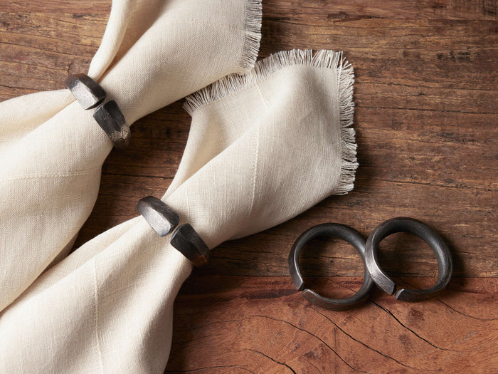 Two folded napkins secured with blackened Arber napkin rings, placed on a wooden surface alongside two additional rings.