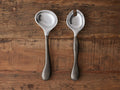 Arber Organic Serving Spoons with polished tops and distressed handles, set of two on textured wooden surface.