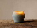 Arhaus Elements Candle in a faceted, frosted glass holder, glowing warmly from light gray-green to yellow on a wood surface.