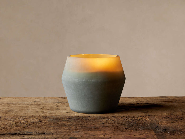 Arhaus Elements Candle in a faceted, frosted glass holder, glowing warmly from light gray-green to yellow on a wood surface.