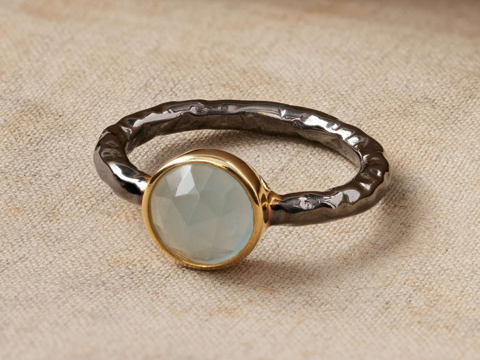 Arne Aquamarine Ring: Oval light blue aquamarine in gold vermeil, atop a dark, hammered smoky pewter band, on textured fabric.