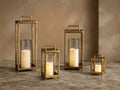 Four glowing Arren Footed Lanterns, brass finish frames, clear glass, varying sizes, on a textured stone floor.