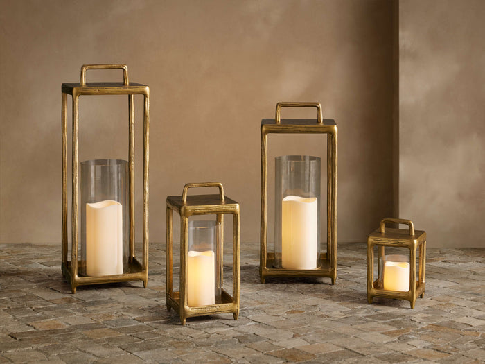 Four glowing Arren Footed Lanterns, brass finish frames, clear glass, varying sizes, on a textured stone floor.